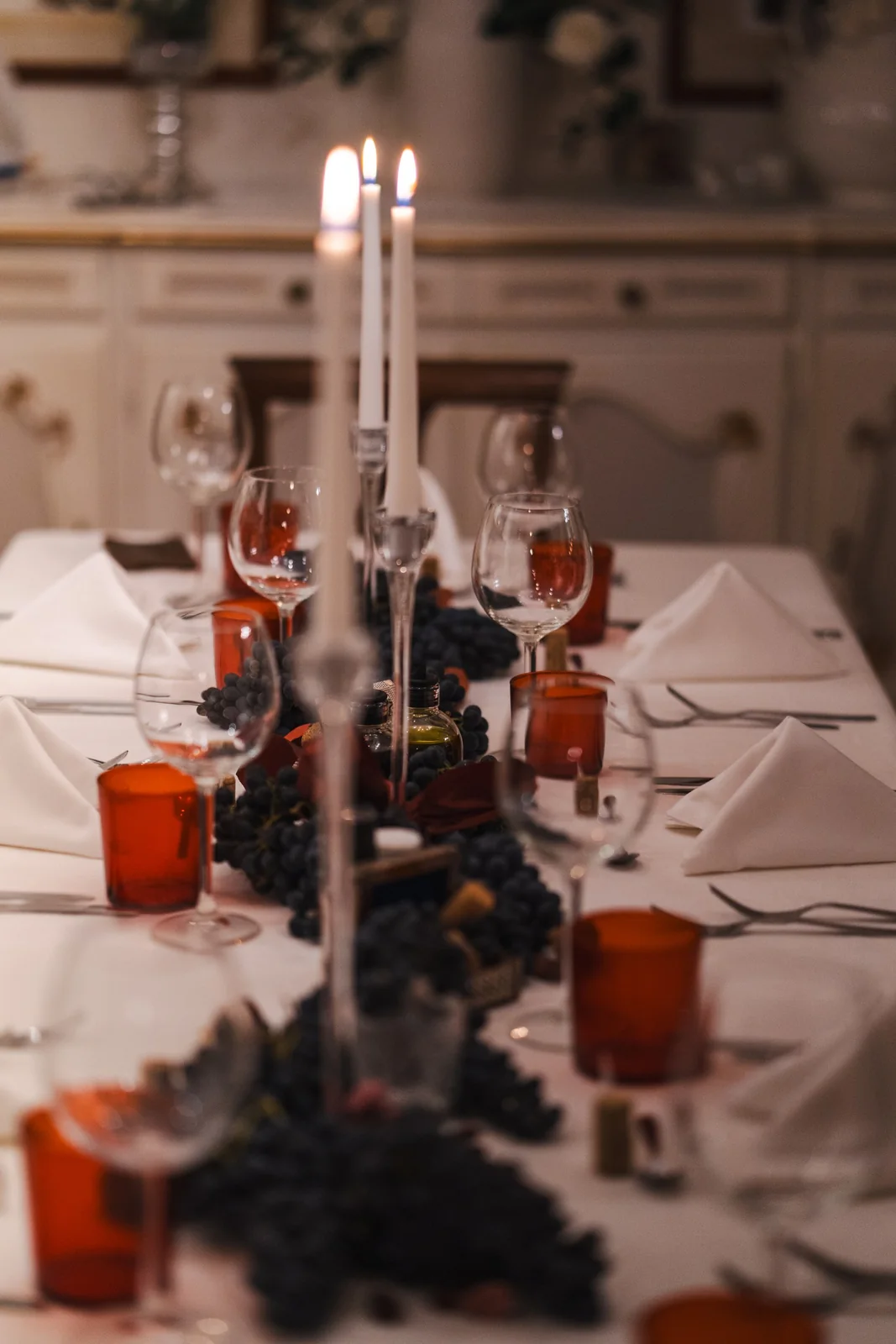 A white celebration table with deep red candles, glasses and grapes